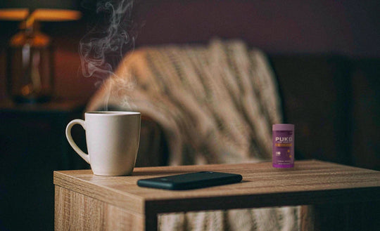 A mug of ashwagandha tea, a phone for scale and the PUKO Unwind + Sleep bottle, which is smaller than the phone, on a wooden side table in a warm dim room at night.